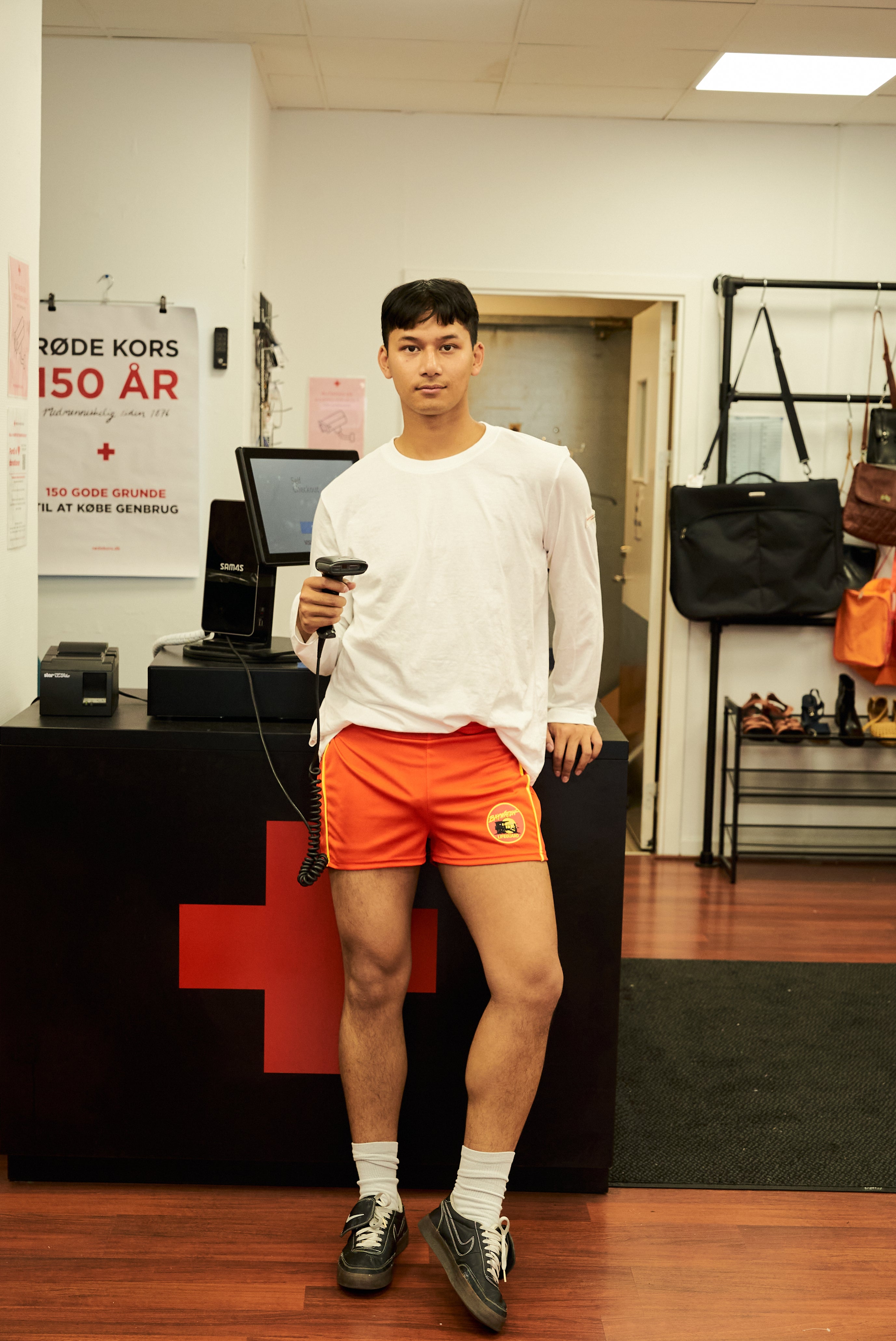 Mandlig model i hvid Boom Langærmet T-shirt og orange shorts – stående ved kassen i Røde Kors genbrugsbutik med stregkodescanner i hånden