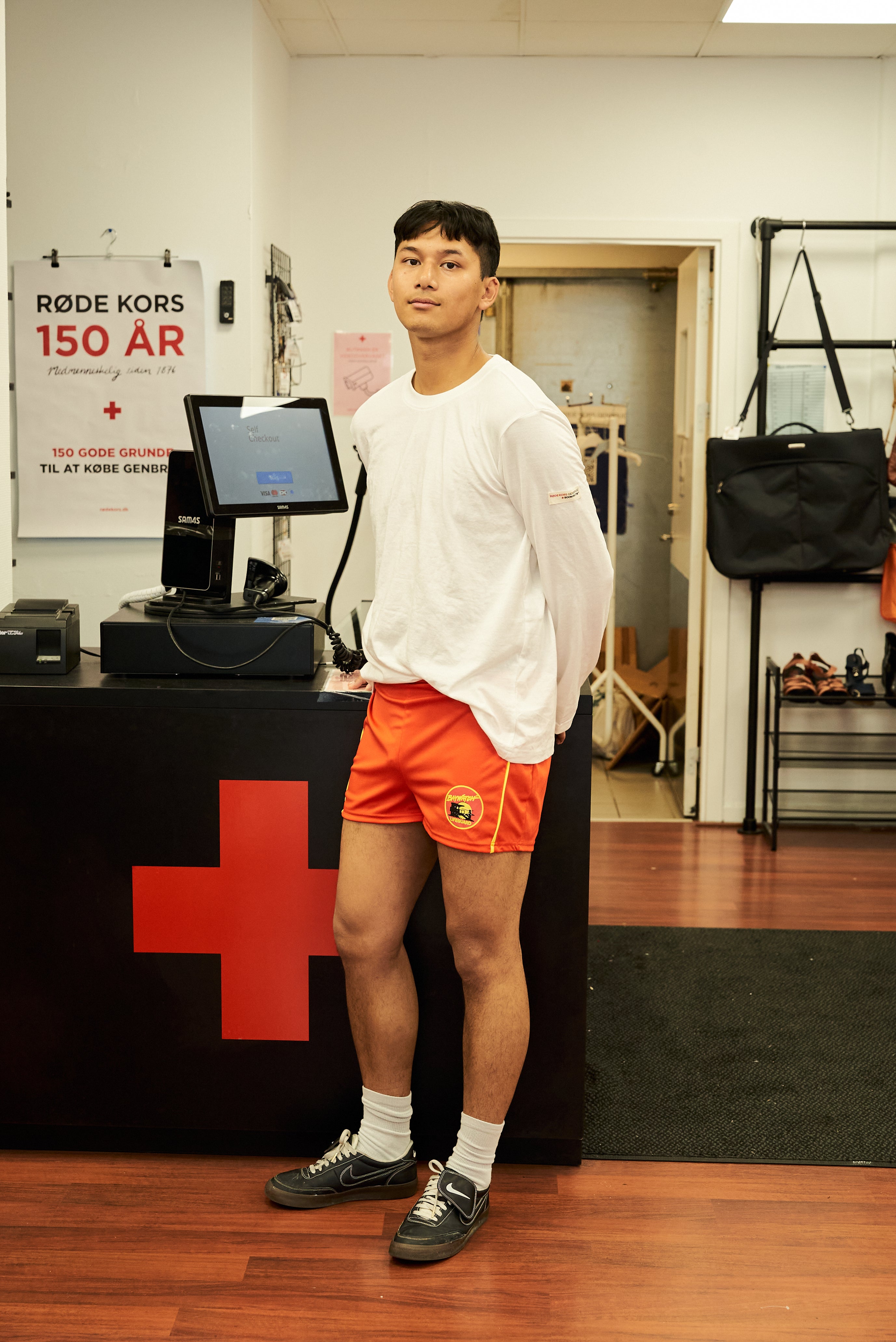 Mandlig model iført hvid Boom Langærmet T-shirt og orange shorts – stående ved kassen i Røde Kors genbrugsbutik